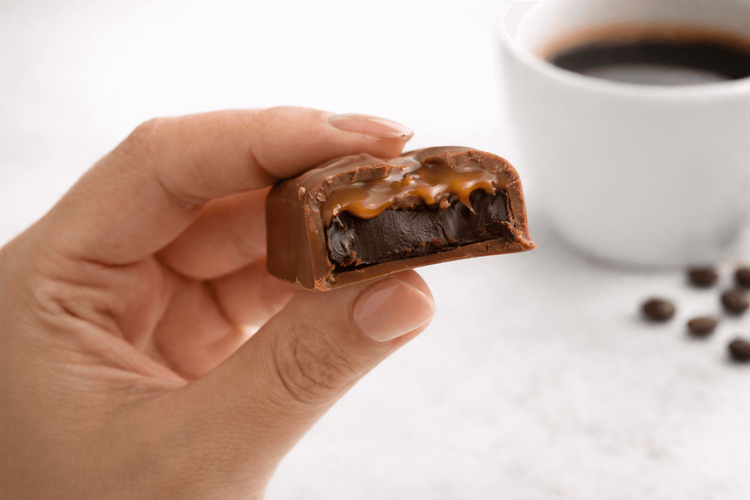 Dolgulu bar çikolata el ile tutulmuş, çikolata içi ve kahve fincanı arka planda.